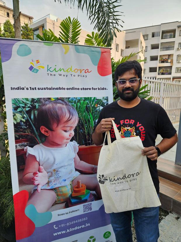 Man holding a 'kindora' tote bag with a promotional banner in the background.