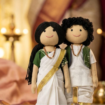Two handmade fabric dolls dressed in traditional Indian attire, standing together against a warm, softly blurred background with warm lights.