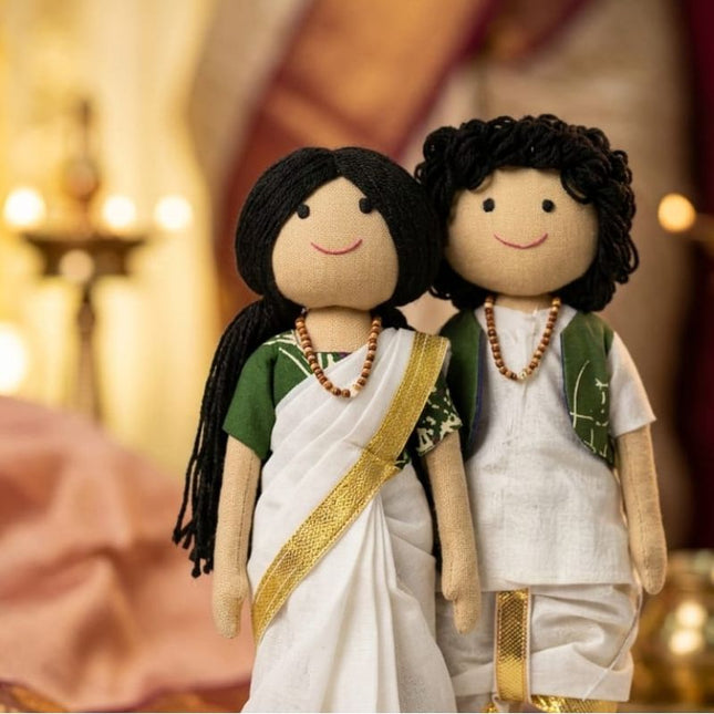 Two handmade fabric dolls dressed in traditional Indian attire, standing together against a warm, softly blurred background with warm lights.