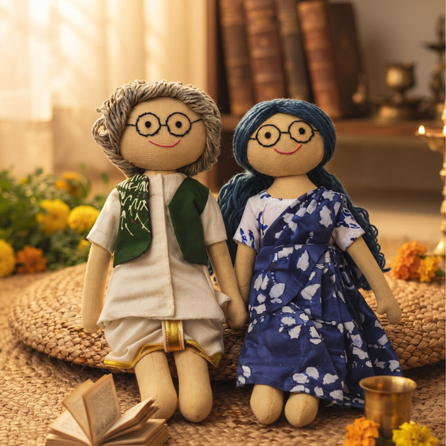 Two handmade wooden dolls with fabric clothing and glasses, posed together on a woven mat in a warm, softly lit indoor setting with books and flowers in the background.