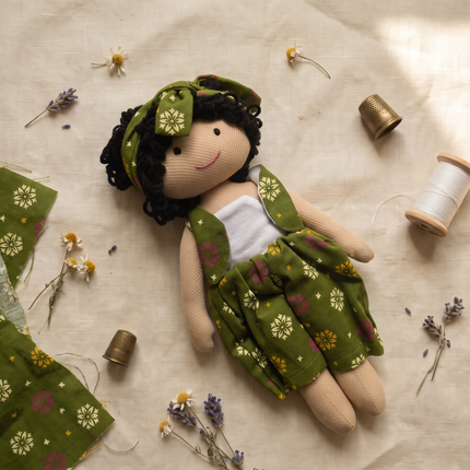 A handmade fabric doll with a green floral dress, matching headband, and black yarn hair, styled on a beige linen background with sewing spools, lavender, and fabric pieces.