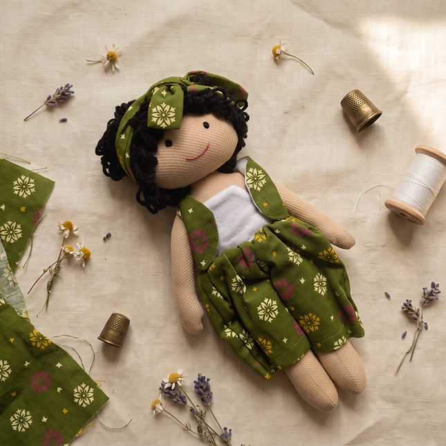 A handmade fabric doll with a green floral dress, matching headband, and black yarn hair, styled on a beige linen background with sewing spools, lavender, and fabric pieces.