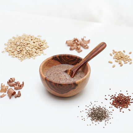 Wooden bowl with a wooden spoon containing a brown spice mix, surrounded by various seeds and spices on a white background.